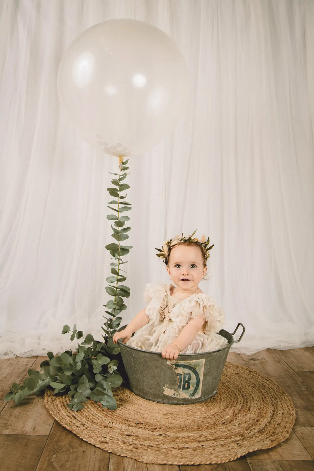 petite fille en studio de photographie dans une bassine en inox avec un très gros ballon et de l'eucalyptus et une couronne de fleurs séchées qui regarde l'objectif
