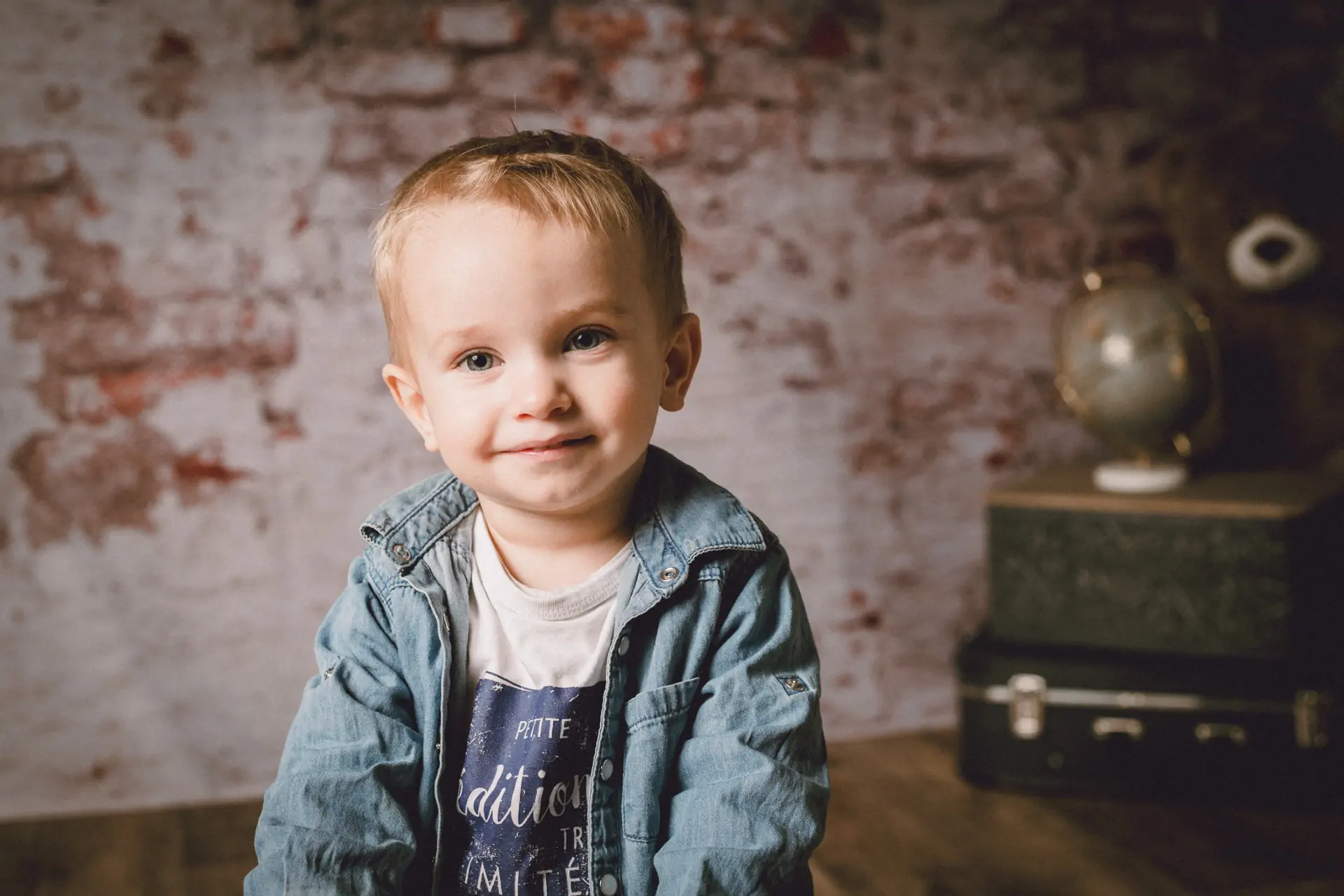 petit garçon dans un studio de photographie il regarde l'objectif du photographe il porte une chemise en jean et le décor en fond est un mur de briques