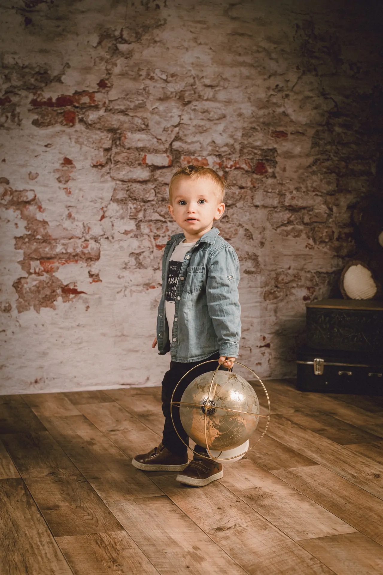 petit garçon debout tenant une mappemonde dans la main dans un studio de photographie il regarde l'objectif du photographe il porte une chemise en jean et le décor en fond est un mur de briques