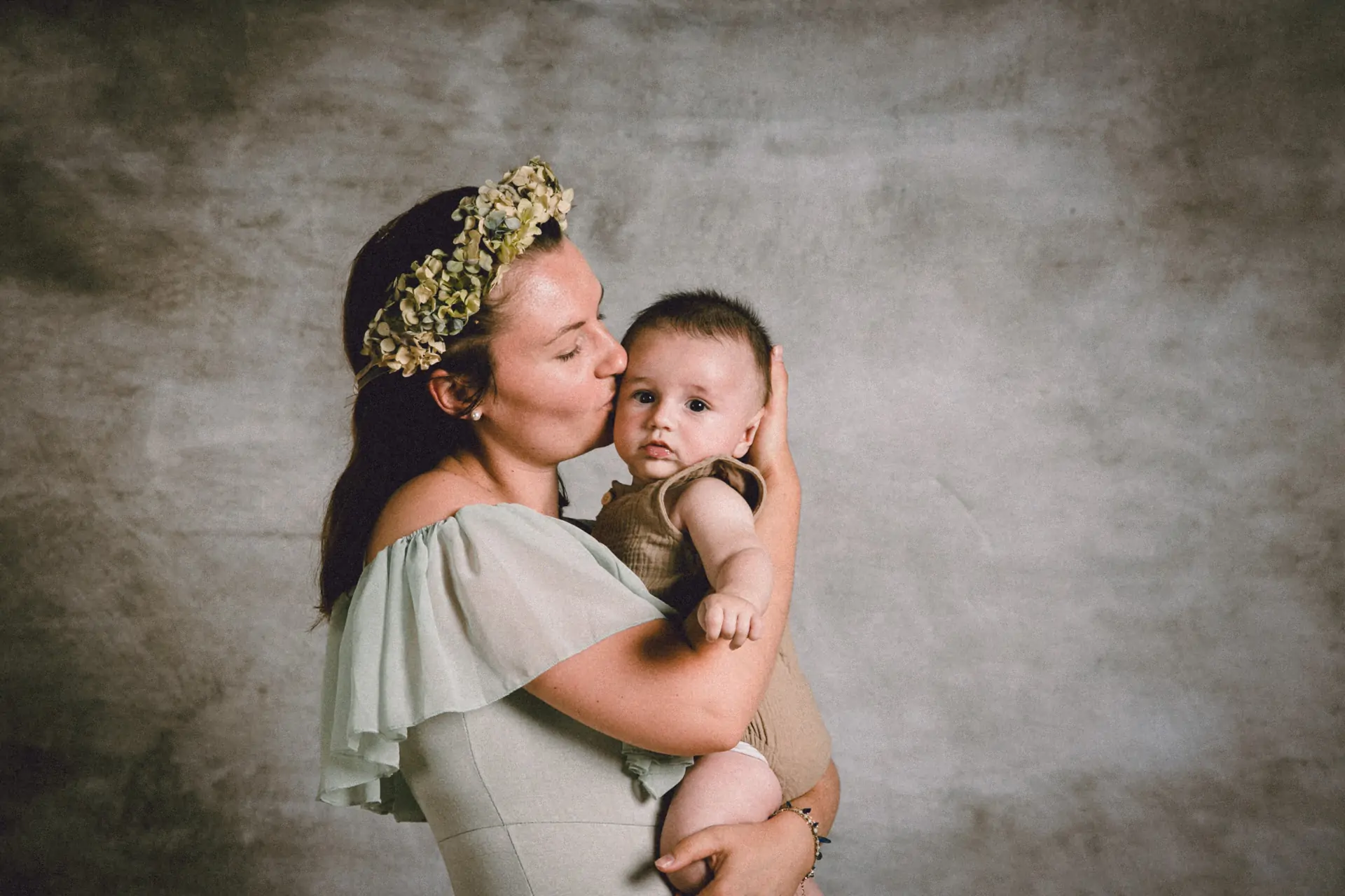 une jeune maman en robe menthe qui fait un bisou à son bébé en studio de photographie avec une couronne de fleurs séchées