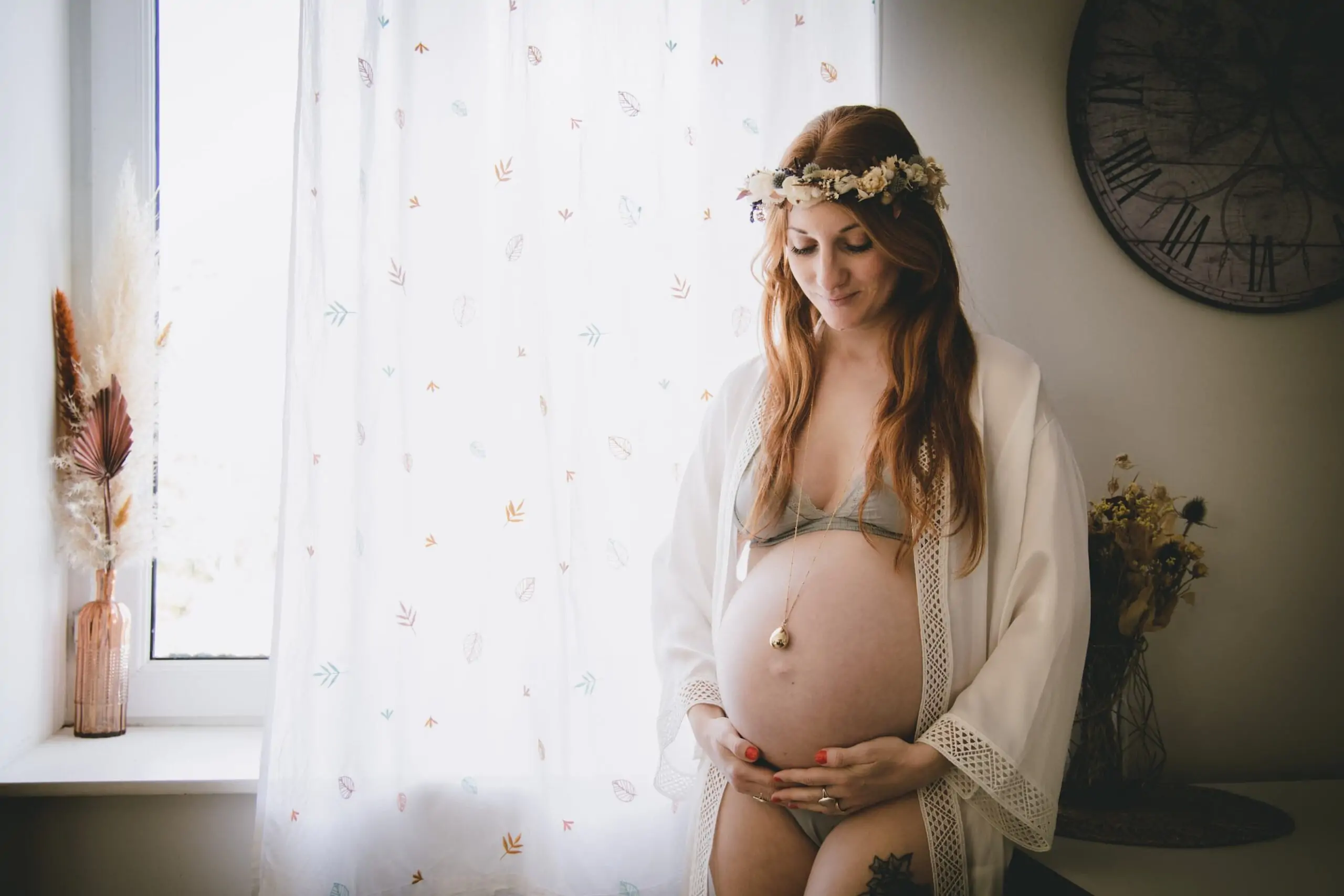 ventre d'une maman qui attend un bébé devant la fenêtre en lingerie et peignoir avec un bola et une couronne de fleurs séchées